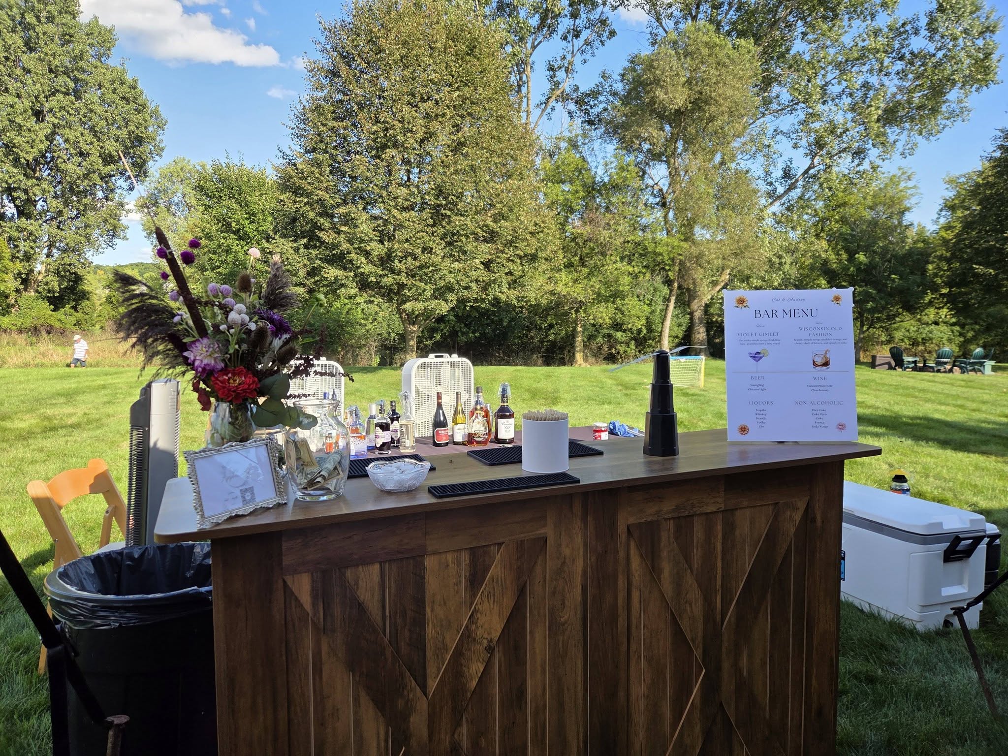 A rustic wooden bar on a lawn with bottles, flowers and a bar menu